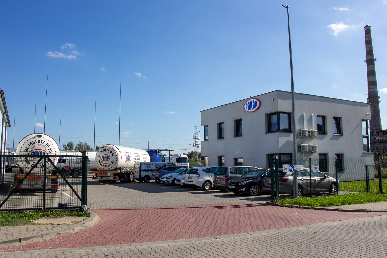 Wjazd na plac PAKAR: za ogrodzeniem z bramą autocysterny, po prawej budynek biurowy z logo PAKAR i parking